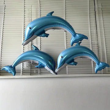 Three large dolphin balloons arranged in an indoor space with white blinds, creating a fun sea life atmosphere for celebrations.
