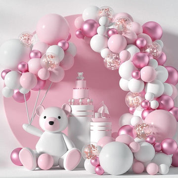 Pink and white balloon garland arch with a teddy bear, cake, and pink balloons for a baby shower or birthday celebration.